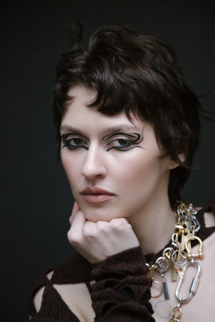 Model with avant-garde graphic eyeliner and soft nude lips, showcasing bold, artistic makeup with intricate black line designs around the eyes, complemented by natural skin and minimalistic lips. The edgy look is enhanced with short hair and layered jewelry