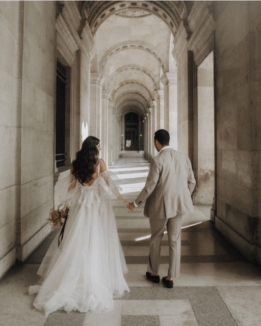 Bride and groom walking hand in hand through a classic Parisian archway, with the bride wearing a flowing gown and soft romantic waves in her hair, showcasing the elegance of a timeless wedding moment.