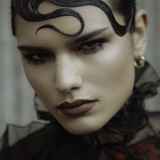 Model with avant-garde makeup featuring deep bordeaux lips, bold graphic eyeliner, and slicked-back hair with intricate finger waves, creating a dramatic and artistic high-fashion look
