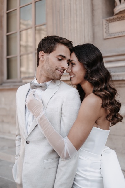 Bride in a sleek satin gown and groom in a light suit share a romantic moment during their Paris wedding, with the bride's hair styled in soft waves and her elegant makeup enhancing the natural beauty of the scene