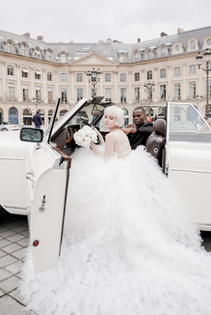 Bride in a voluminous white gown sitting in a vintage car at Place Vendôme, Paris, showcasing chic bridal makeup and a luxurious wedding look.